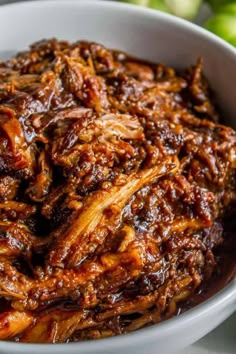 a white bowl filled with cooked meat and sauce on top of a table next to an apple