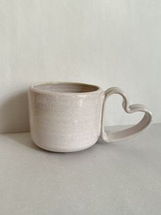 a white coffee cup with a heart shaped handle on the side, sitting in front of a plain background