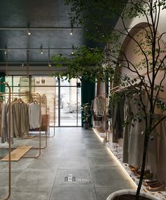 the inside of a clothing store with lots of clothes on racks and trees in pots