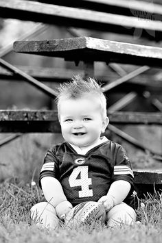 a little boy sitting in the grass with a football on his feet and smiling at the camera