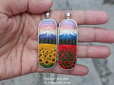 a hand holding two pendants with flowers on them in front of a mountain range