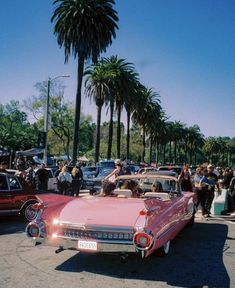 an old pink car parked in a parking lot with palm trees and people standing around