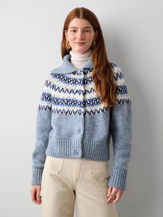 a woman standing in front of a white wall wearing a blue sweater and tan pants