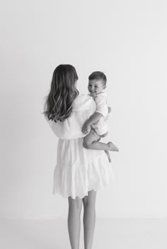 a woman holding a baby in her arms while standing against a white wall and looking at the camera