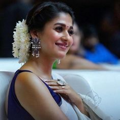 a woman sitting on a white couch wearing large earrings and a blue dress with flowers in her hair