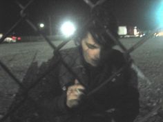 a man sitting behind a chain link fence looking at his cell phone in the dark