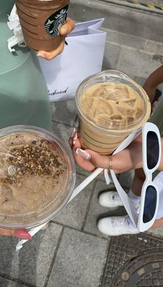 two people are holding drinks and plates with food in them on the sidewalk near some trash cans