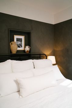 a bed with white linens and pillows in a dark room next to a painting on the wall