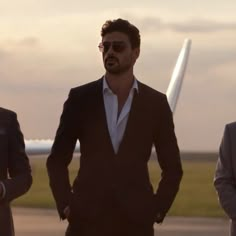 three men in suits standing next to each other on an airport tarmac at sunset