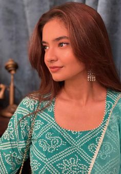 a woman with red hair wearing a green blouse and earrings standing in front of a blue curtain