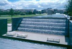 a large water fountain in the middle of a park with benches around it and buildings in the background