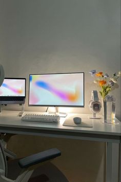 two computer monitors sitting on top of a desk next to a vase with flowers in it