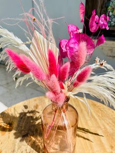 pink flowers in a glass vase sitting on a wooden table with white and pink feathers