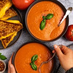 two bowls of tomato soup with grilled cheese on the side and tomatoes, basil leaves