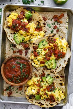 breakfast tacos with eggs, avocado and salsa on a baking sheet next to a bowl of salsa