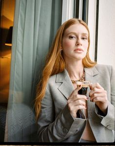 a woman with long red hair is holding a glass in her hand and looking out the window