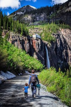 a man and child walking down a dirt road next to a waterfall