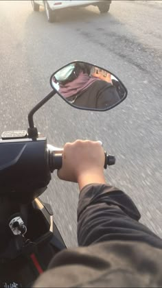 a person riding a motorcycle on a street with a car in the background and someone's reflection in the rear view mirror