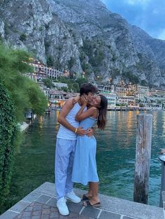 a man and woman kissing in front of a body of water with mountains in the background