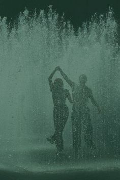 two people standing in front of a fountain with water shooting up from the top and behind them