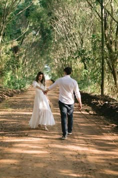 a man and woman walking down a dirt road in front of trees on both sides