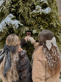 two women standing next to each other in front of a christmas tree with snow on it