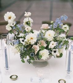 an arrangement of flowers in a vase on a white table cloth with candles and place settings