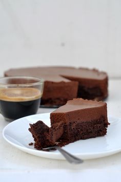 a piece of chocolate cake on a white plate with a cup of coffee in the background