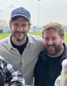 three men standing next to each other in front of a baseball field with one man wearing a hat