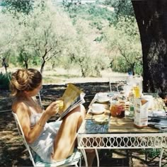 a woman sitting at a table reading a book in the shade of an olive tree