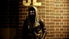 a man wearing a black mask and leather jacket standing in front of a brick wall