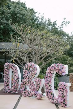 the letters are decorated with pink flowers in front of a tree and shrub behind it