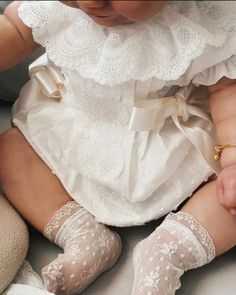 a baby in white dress and lace socks sitting next to a teddy bear