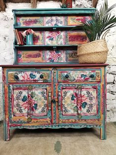 an old painted dresser with flowers and plants on top, next to a brick wall