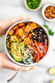 two hands holding a bowl filled with different types of vegetables and black beans, avocado