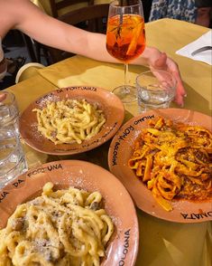 three plates of pasta and wine on a table with two people holding glasses in the background