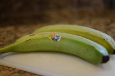 two green bananas sitting on top of a white cutting board next to a brown counter