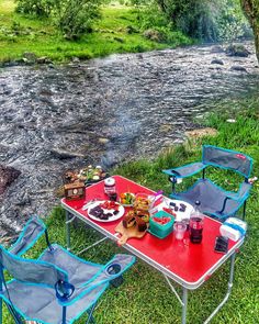 two chairs and a table with food on it near a stream in the grass next to some trees