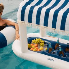 a woman sitting on an inflatable raft with drinks and fruit, next to the pool