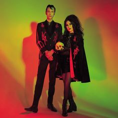 a man and woman standing next to each other in front of a colorful wall holding a skull