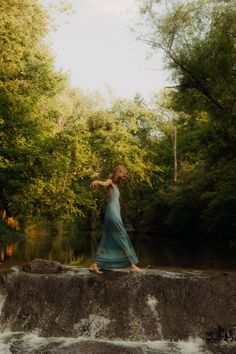 a woman in a blue dress is standing on a rock by the water's edge