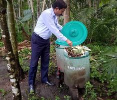 a man standing next to a green trash can filled with dirt and plants in the woods