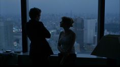 two women standing in front of a window looking out at the cityscape and skyscrapers