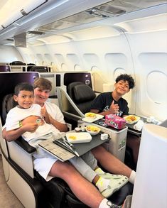 three boys sitting on an airplane with food in their lap top trays and one boy is eating