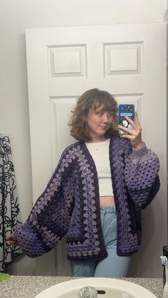 a woman taking a selfie in front of a bathroom mirror wearing a purple crocheted cardigan