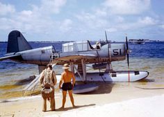 two men standing on the beach next to an airplane in the water with another man looking at it