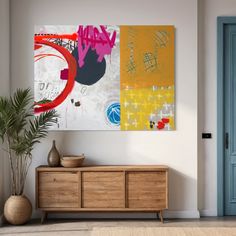 an abstract painting hangs on the wall next to a wooden cabinet and potted plant