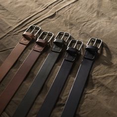 four different belts are lined up on a brown cloth covered bed sheet, one has a metal buckle and the other has a black leather belt