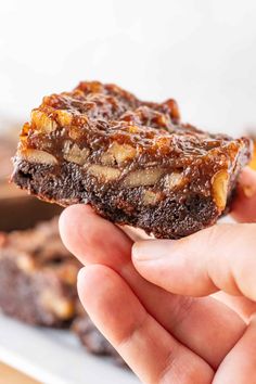 a hand holding up a piece of brownie with nuts on top and chocolate in the background