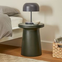 a small table with a lamp on it next to a basket and bed in the background